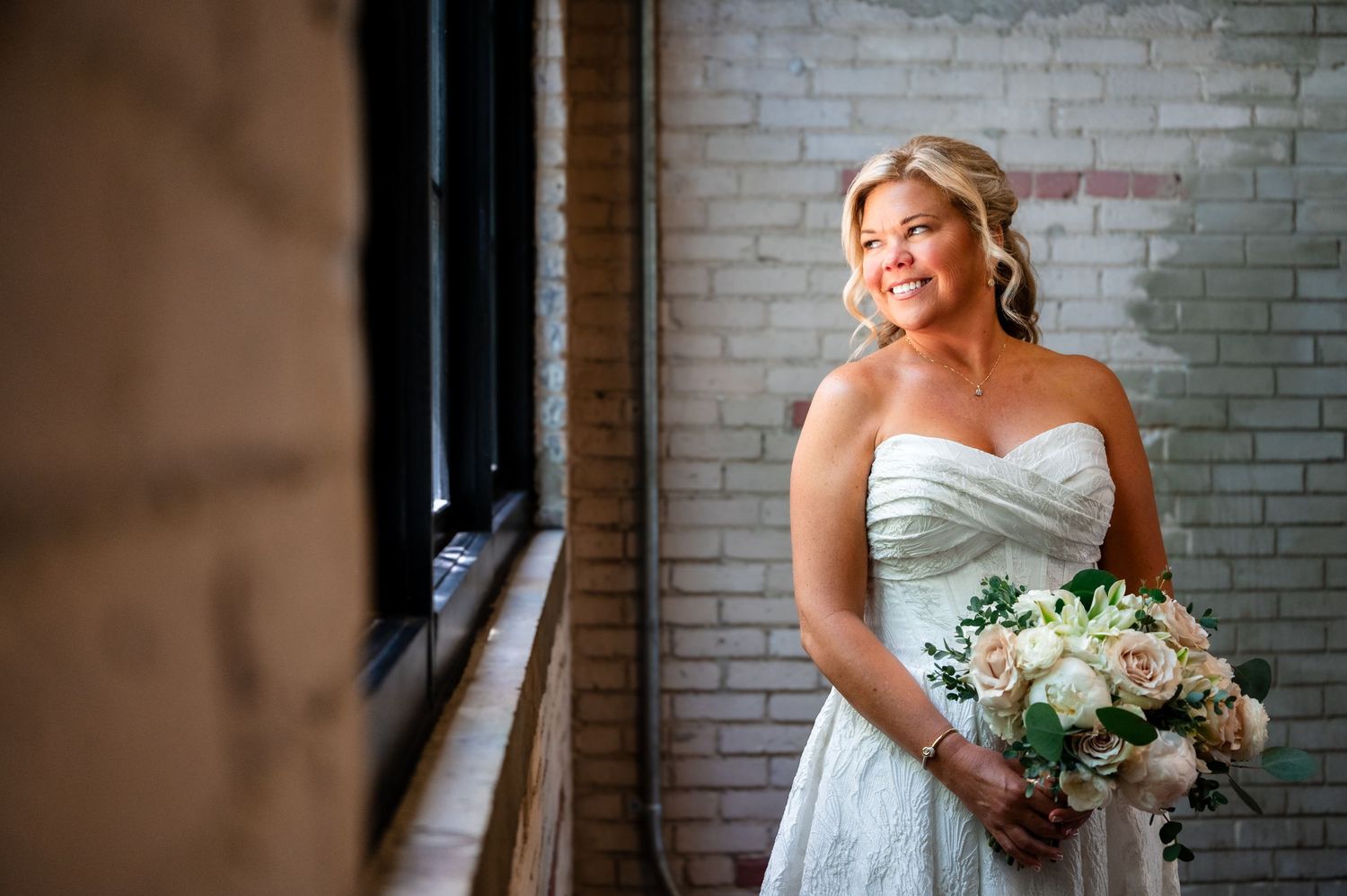 Smiling bride in strapless lace gown holding white bouquet, standing by window with brick wall backdrop.