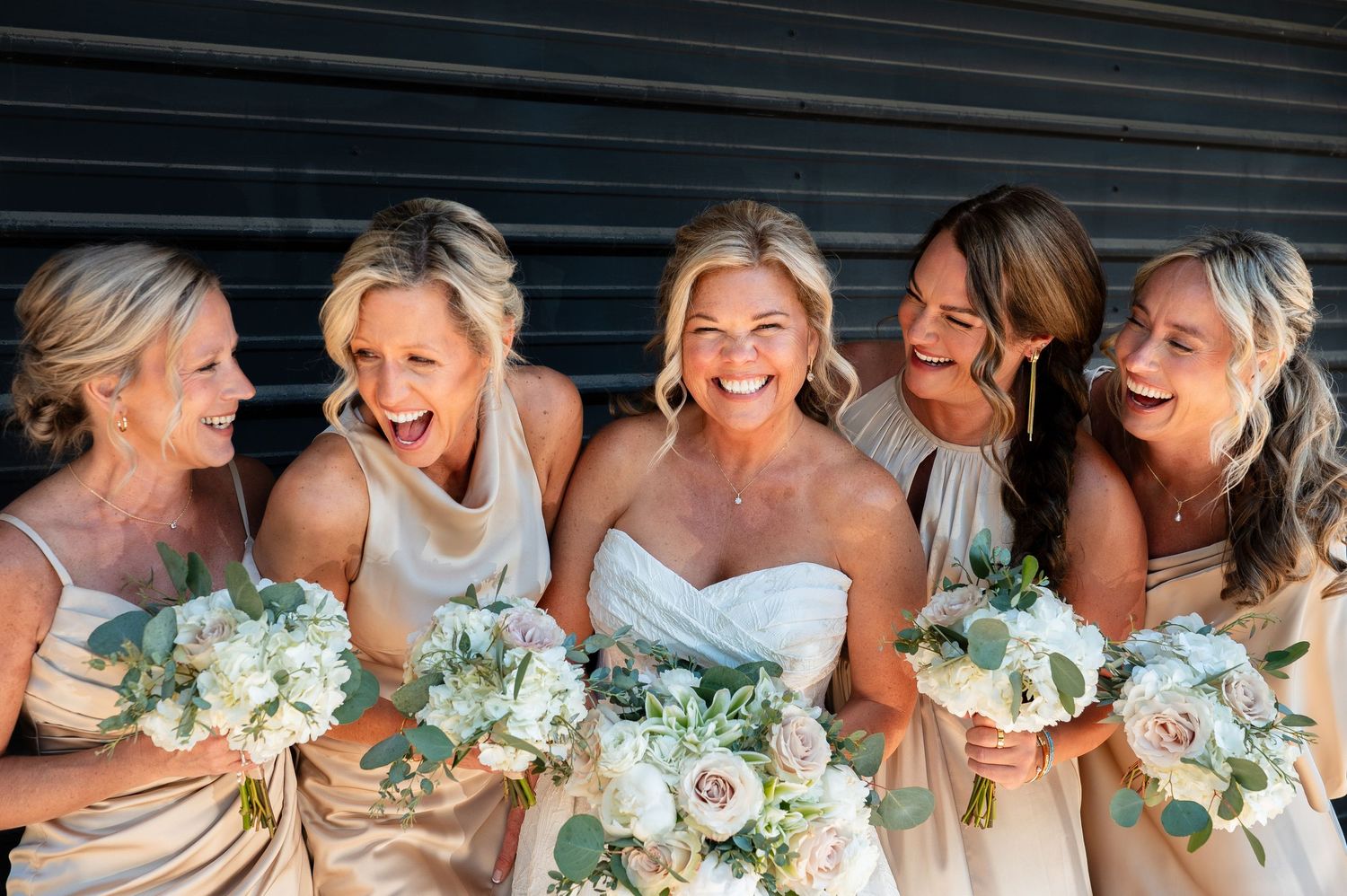 Bride and bridesmaids in champagne dresses laughing together holding white floral bouquets with eucalyptus greenery.