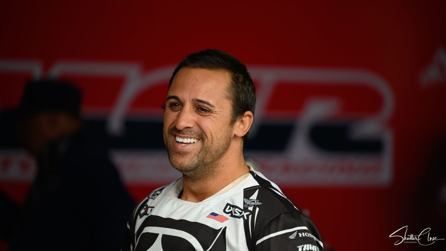 Smiling male motocross rider in black racing gear posing at an indoor motorsport event.