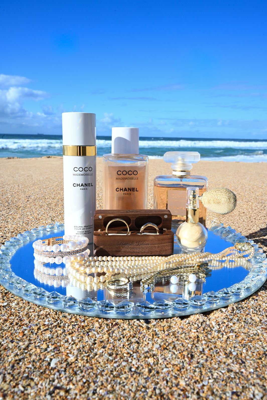 Chanel Coco perfume and body spray on a mirrored tray with pearl jewelry and gold rings on a sunny beach.