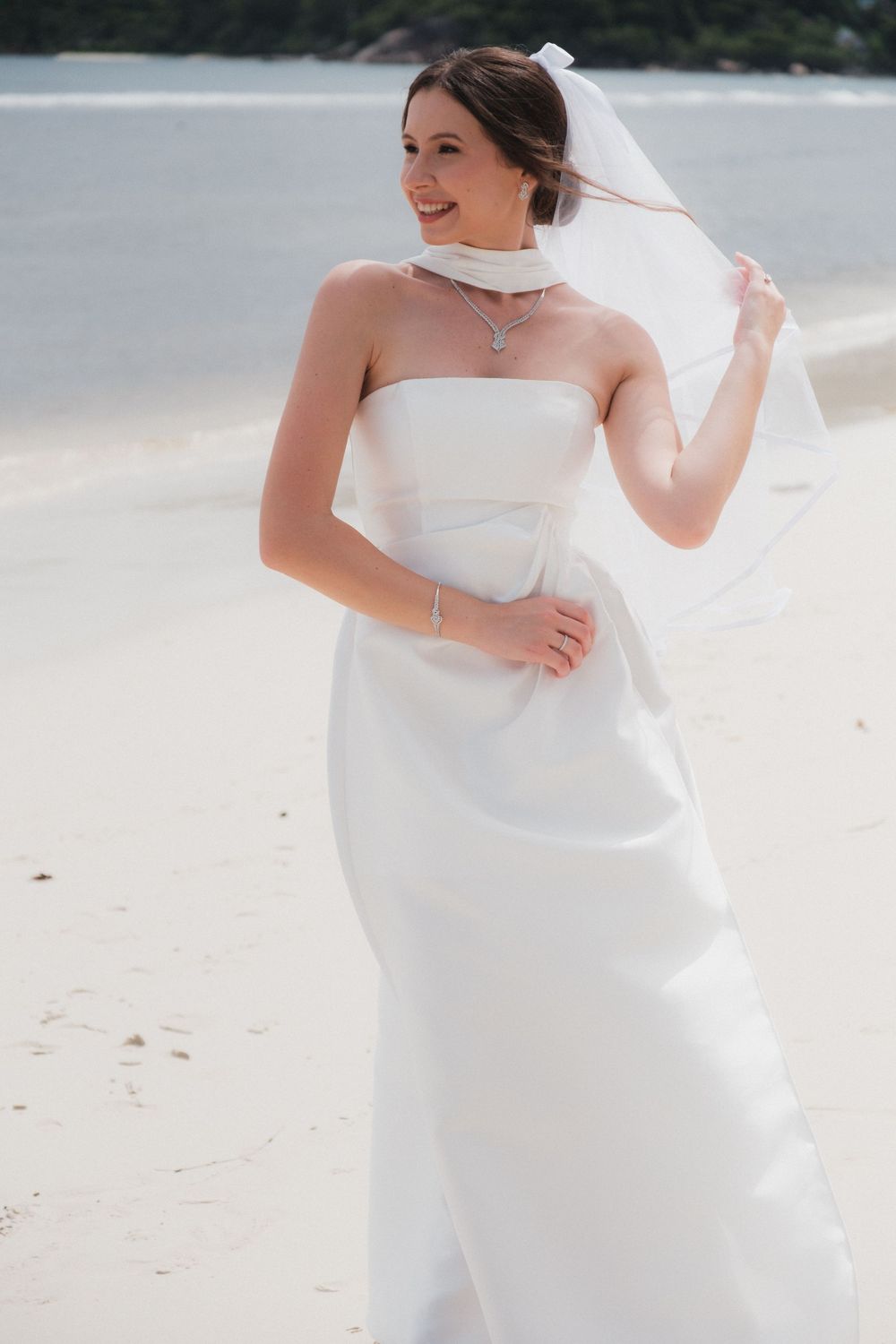 Smiling bride in strapless white gown and veil on sandy beach, holding veil in wind.