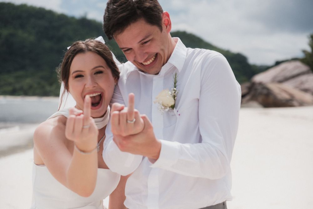 Happy newlyweds showing wedding rings on beach, bride in white dress and groom in white shirt smiling joyfully.