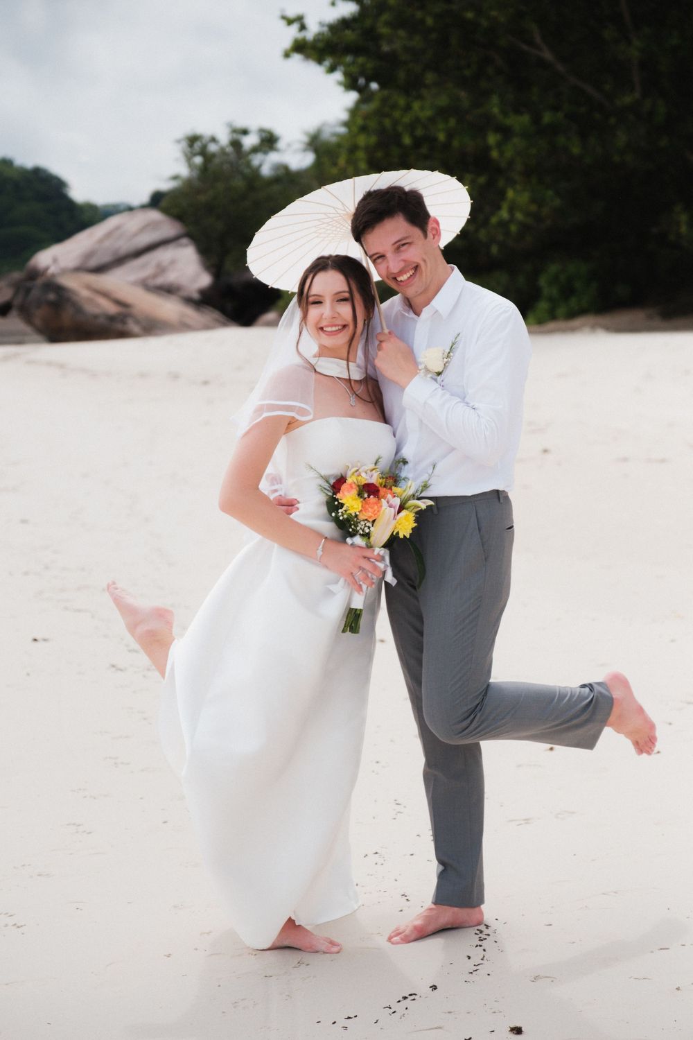 Happy newlyweds pose on a white sand beach, bride holding sunflowers bouquet under white parasol in wedding attire.