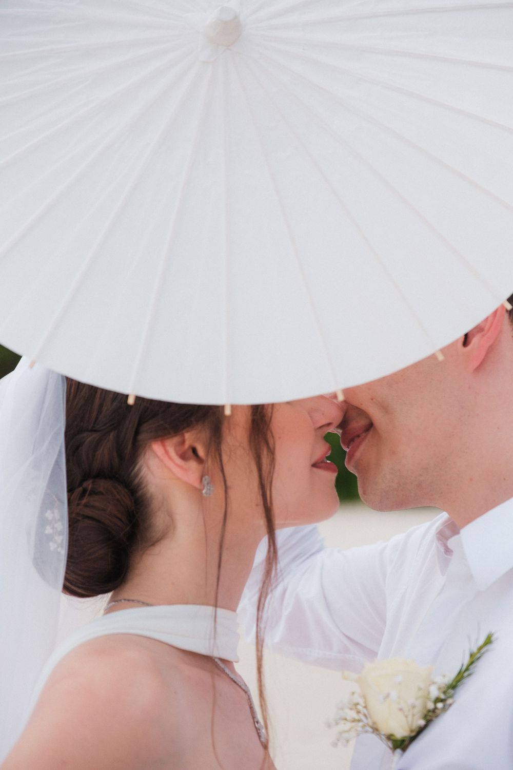 Newlywed couple sharing a romantic kiss under a white paper parasol on their wedding day.