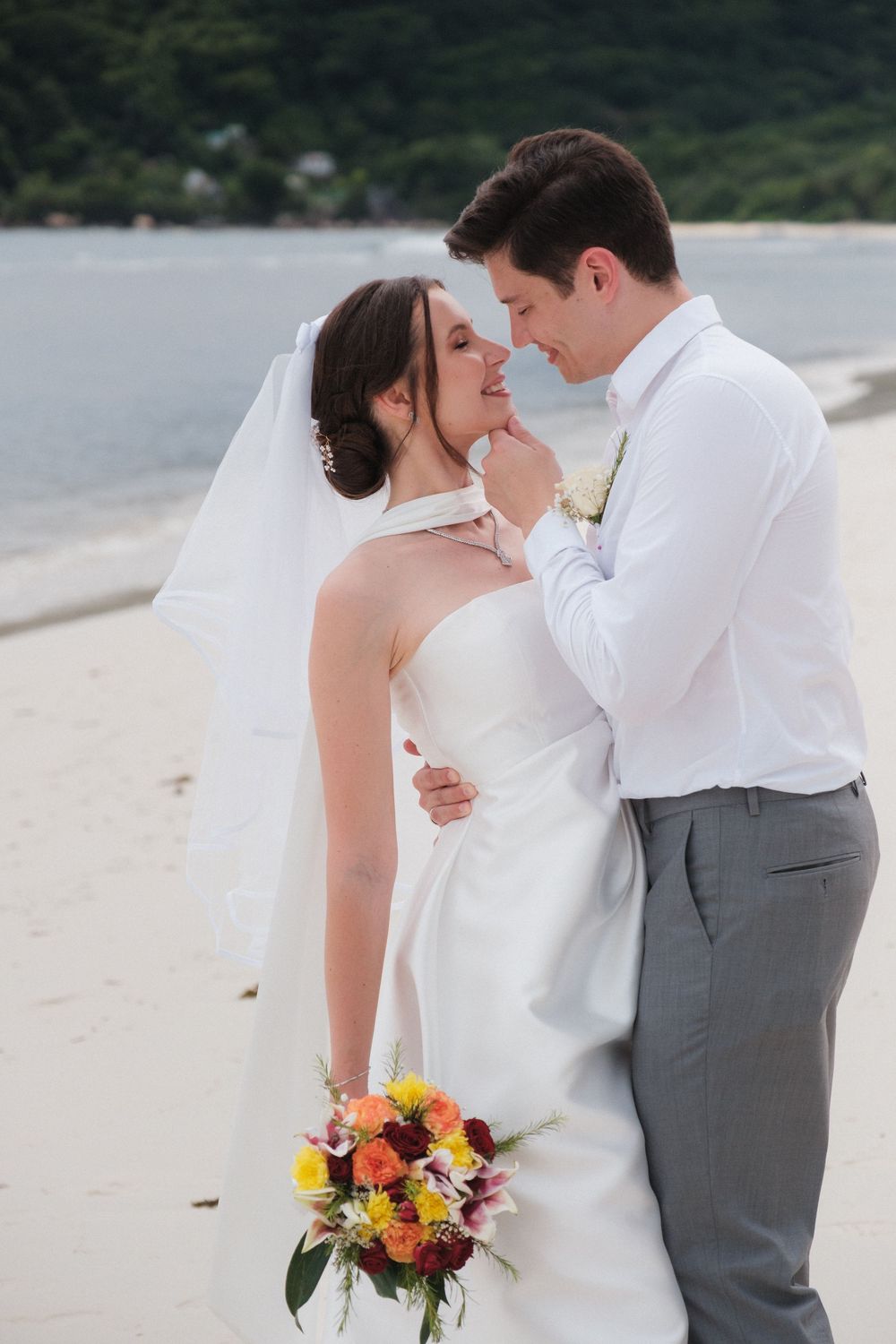 Bride and groom share a romantic moment on a sandy beach, with colorful bouquet and veil blowing in the breeze.