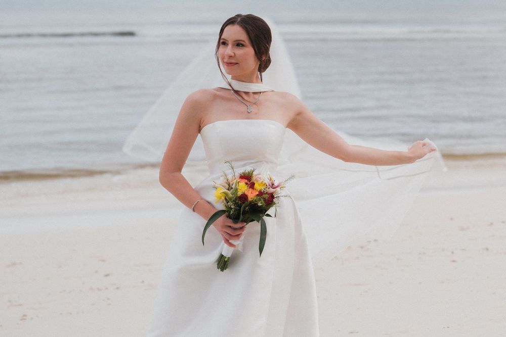 Bride in white strapless gown and veil holds colorful bouquet on sandy beach with ocean backdrop.