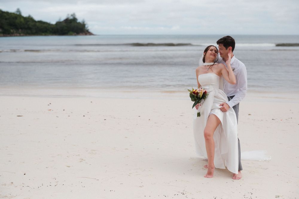 Couple in wedding attire embracing on a white sand beach, bride holding a bouquet with ocean in the background.
