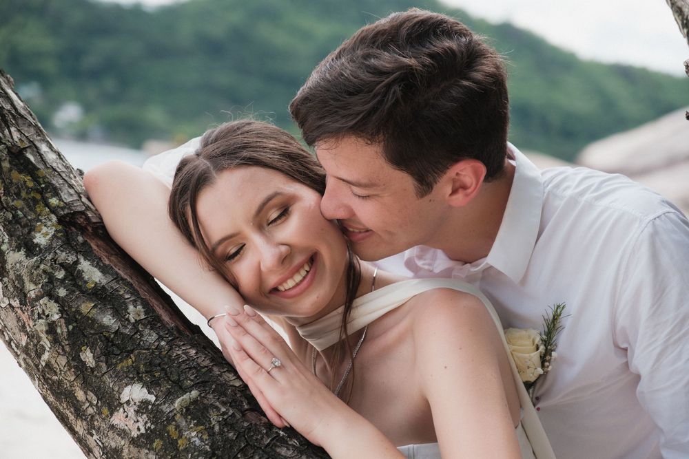 A smiling bride leans against a tropical tree as her groom embraces her tenderly on a beach wedding day.