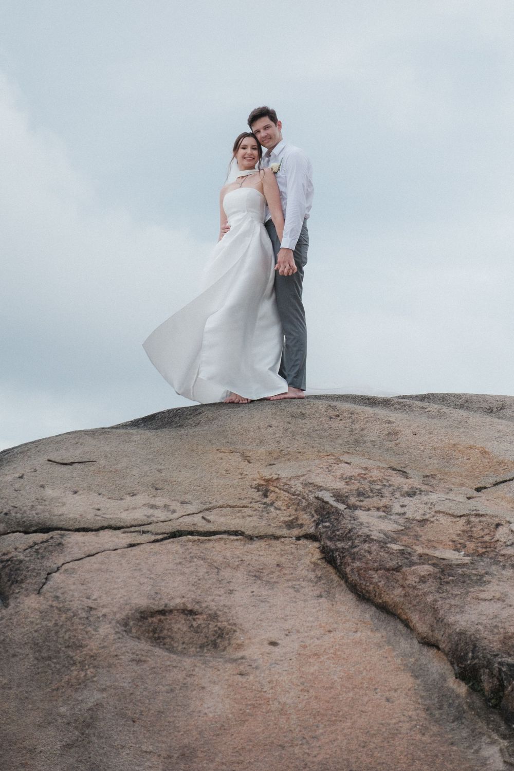 Bride and groom standing atop a rocky cliff, embracing with wedding dress flowing in the wind against a cloudy sky.