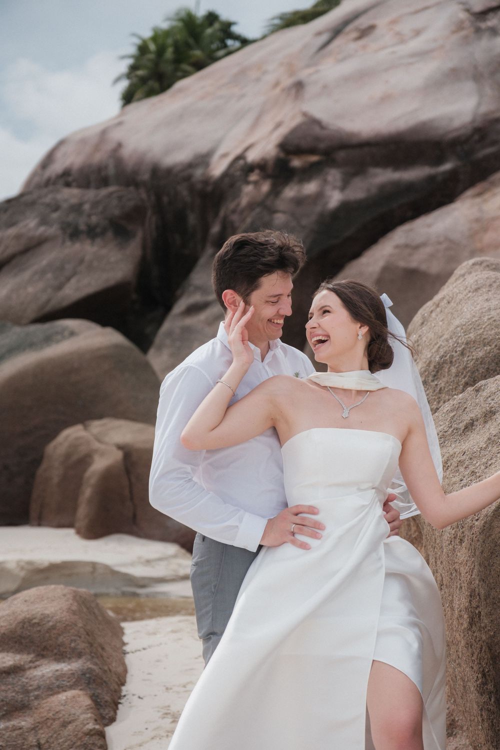 A joyful bride and groom embrace on a rocky beach, the bride in a white strapless gown laughing as they share a romantic moment.