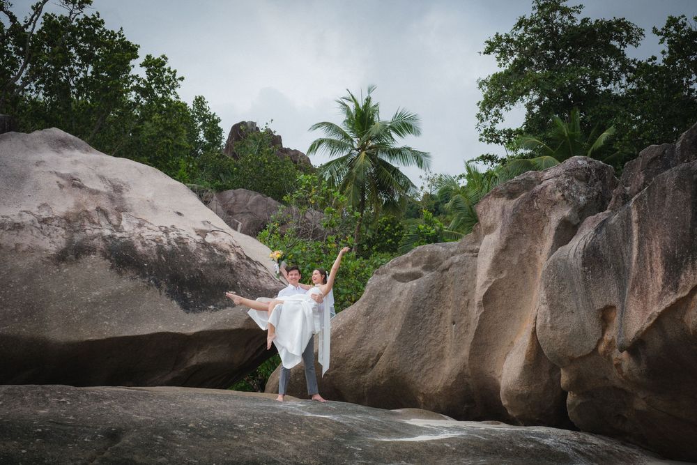 Newlywed couple posing joyfully among large granite boulders surrounded by tropical vegetation.