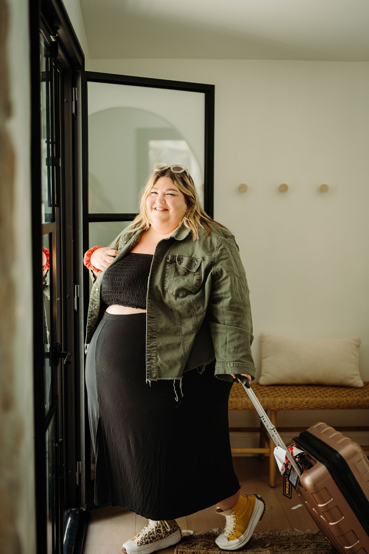 Woman in green jacket and black dress smiling with luggage near a doorway.