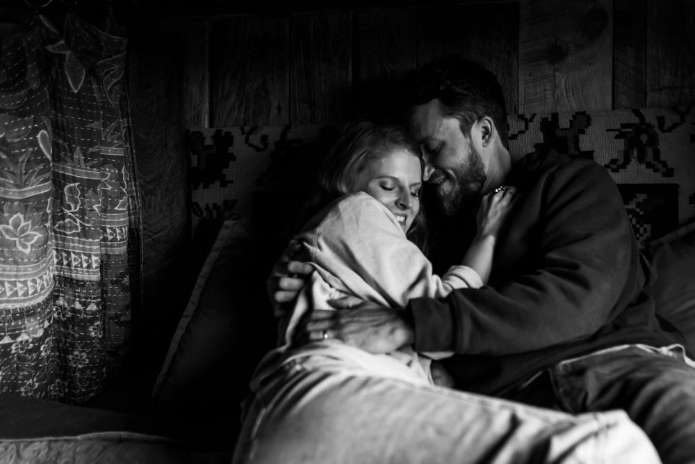 Black and white photo of a couple sharing an intimate embrace, wrapped in a blanket in a cozy rustic setting.
