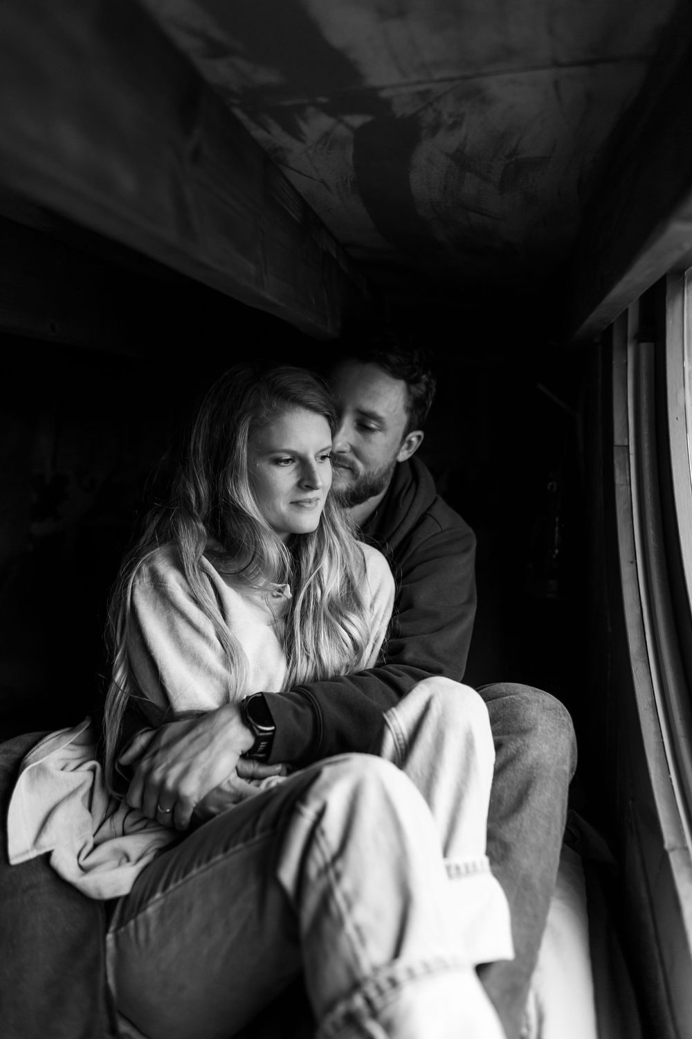 Black and white photo of a couple sitting together by a window, man embracing woman from behind in cozy interior.