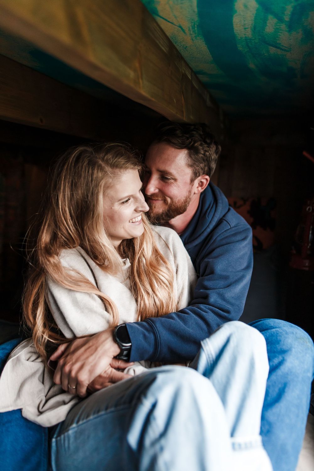 A smiling couple embraces warmly, the woman in a cream sweater and man in a blue hoodie, sitting together in a cozy setting.