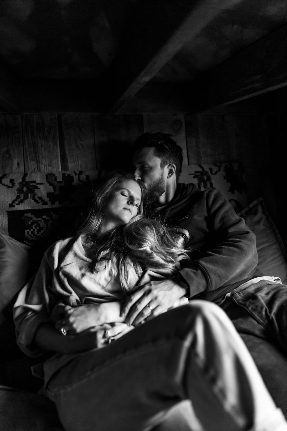 Black and white photo of a couple cuddling intimately on a couch in a dark rustic room.