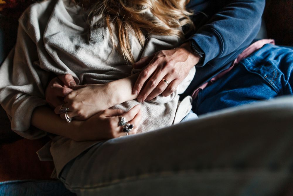 Two people embracing on a couch, hands intertwined, wearing casual clothing in a warm, intimate moment.