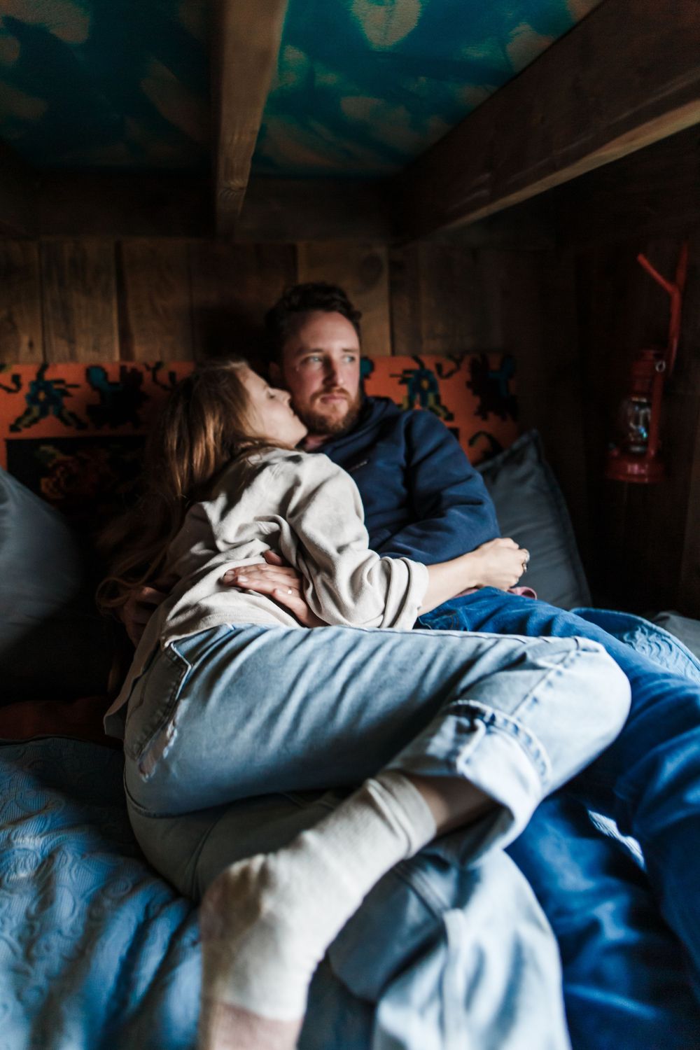 Couple cuddling intimately on a bed in a cozy wooden cabin bunk, woman leaning against man's chest.