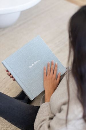 Woman holding a personalized gray linen photo album titled The Andersons while sitting on a woven rug.