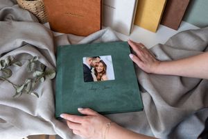 Green velvet wedding photo album held by hands, surrounded by fabric swatches and eucalyptus leaves.