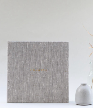 Gray linen wedding photo album with gold foil names Peter & Kate, displayed beside a small ceramic vase.