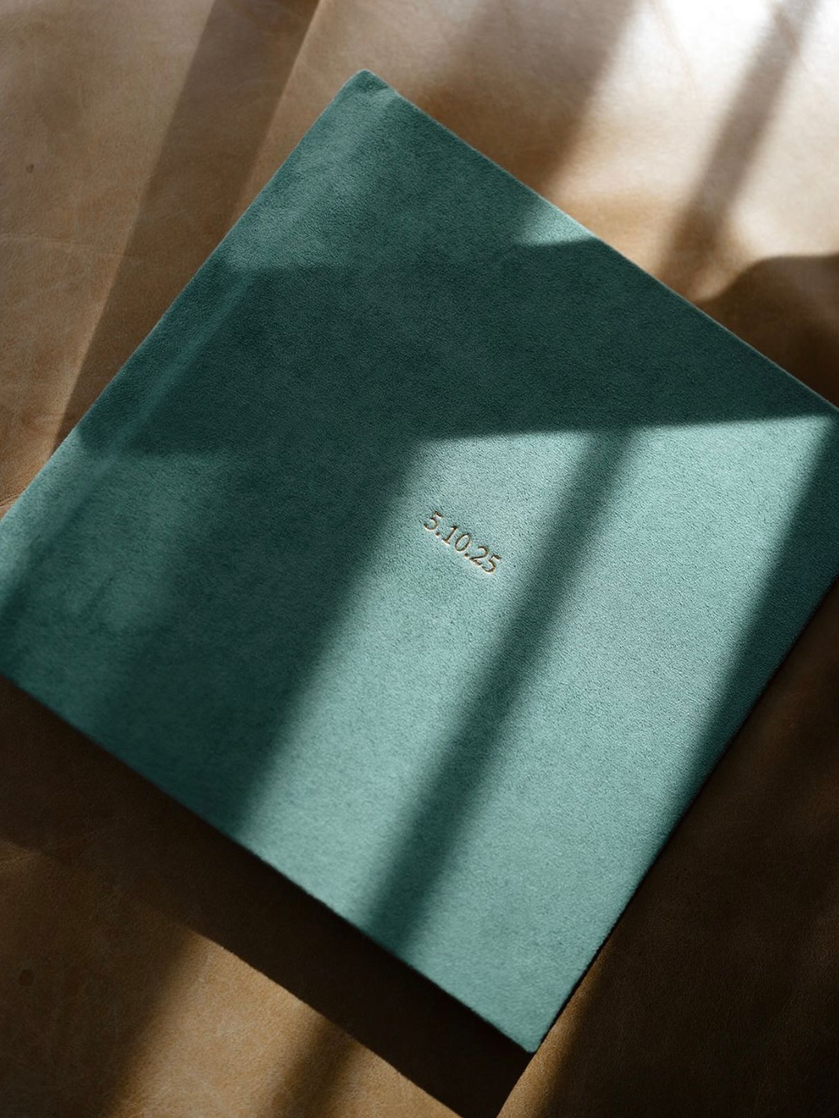 Teal velvet photo album with gold embossed date 4.10.25, placed on a beige surface with soft natural light shadows.
