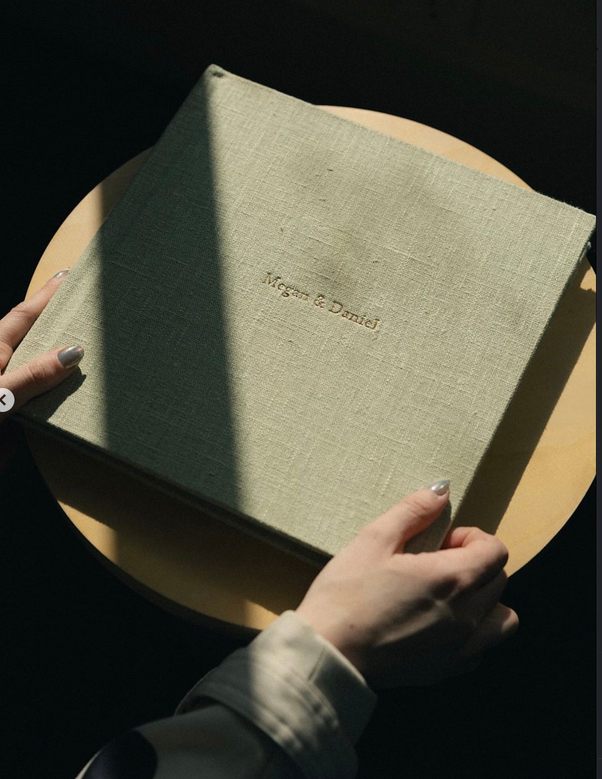 Hands holding a sage green linen-bound wedding photo album embossed with names Megan & Daniel.