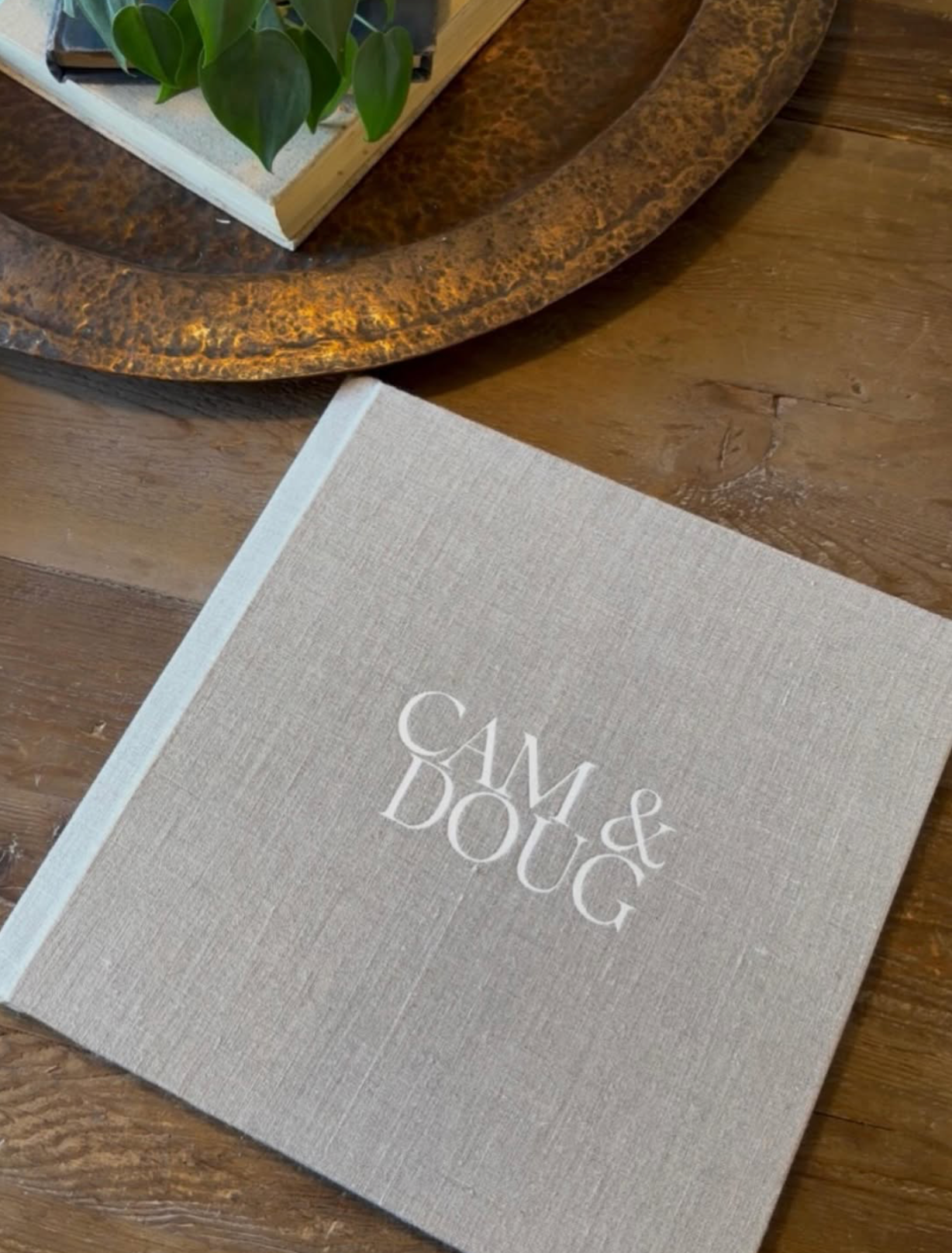 Elegant linen-covered wedding photo book embossed with "Cam & Doug" displayed on a wooden table.