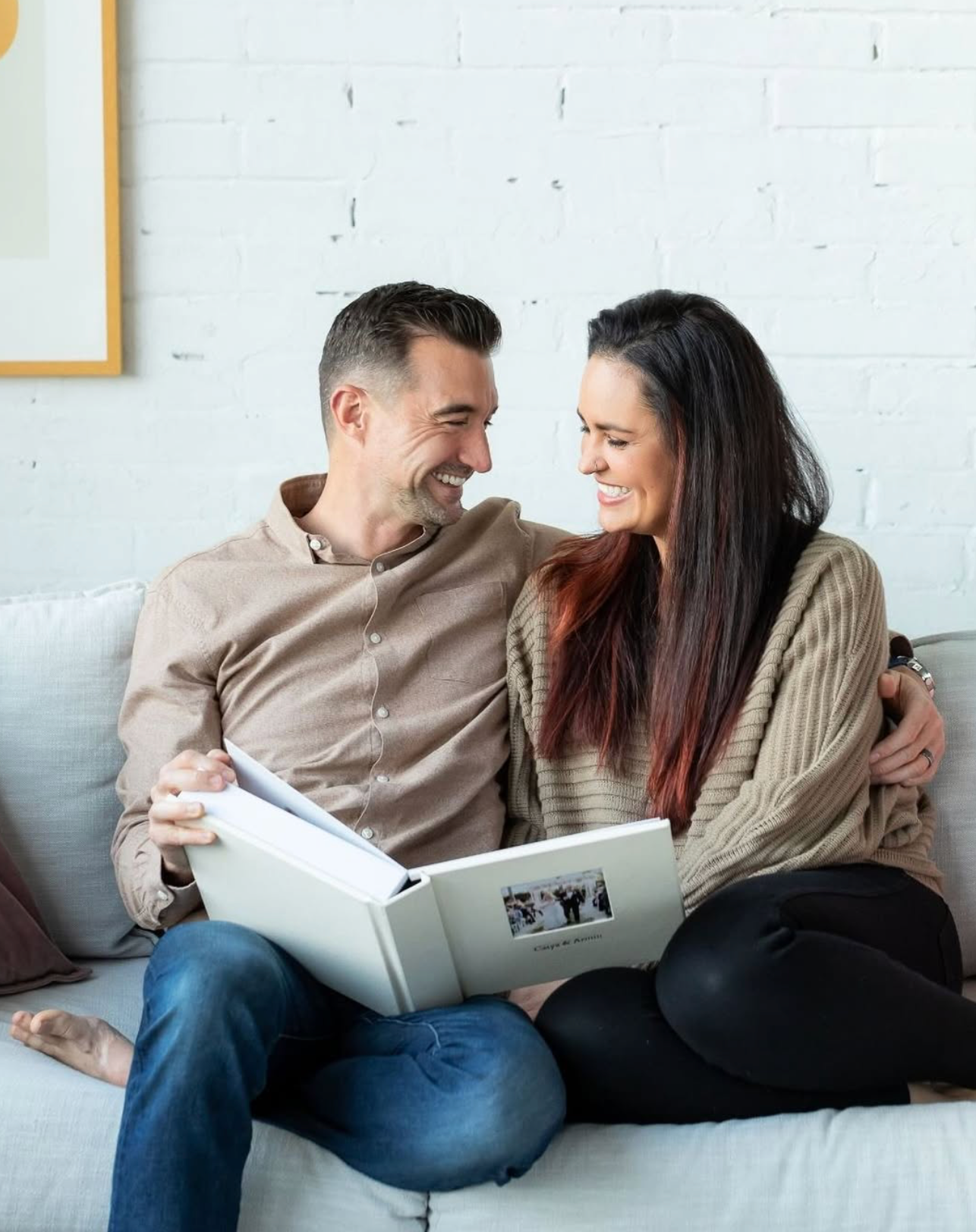 Couple smiling at each other on a couch while looking at a photo album together in a cozy living room.
