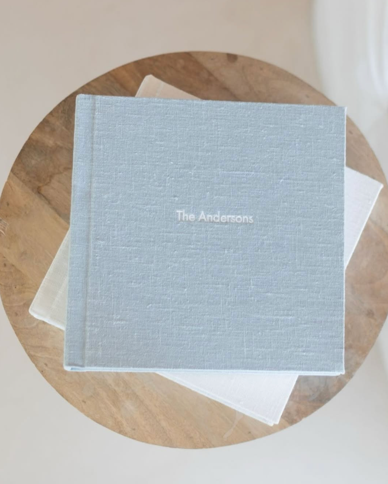 Light blue linen hardcover photo album personalized with "The Andersons" sitting on a round wooden table.