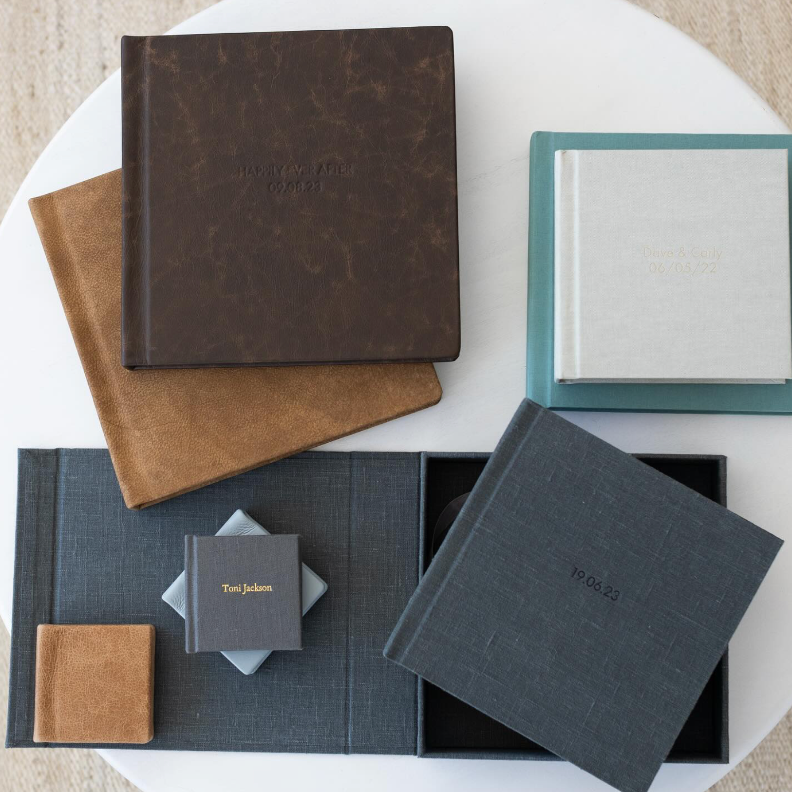 Collection of premium photo albums and keepsake boxes in brown, gray, and teal leather and linen covers displayed on white surface.