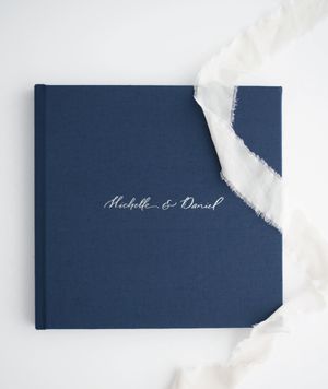 Navy blue linen wedding photo album embossed with cursive names Michelle and Daniel, with white silk ribbon accent.