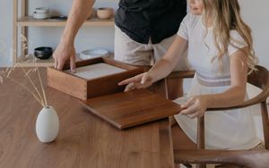 Two people assembling a wooden drawer at a dining table in a modern home interior.