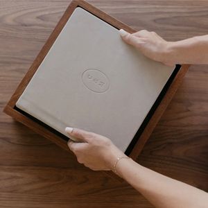 Hands placing a gray embossed book into an open walnut wood box on a dark wooden table.