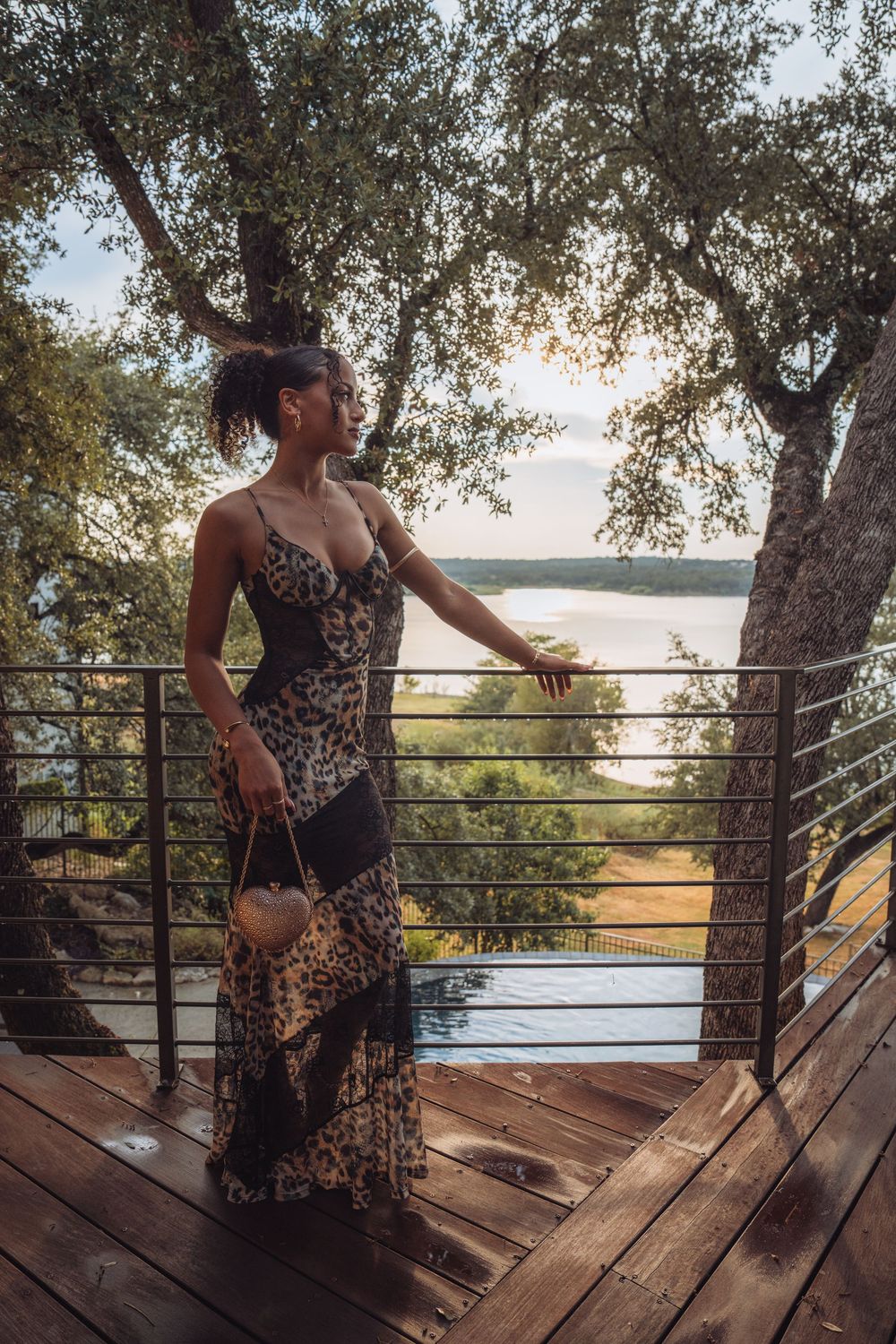 Woman in leopard print maxi dress standing on wooden deck overlooking scenic lake at sunset.