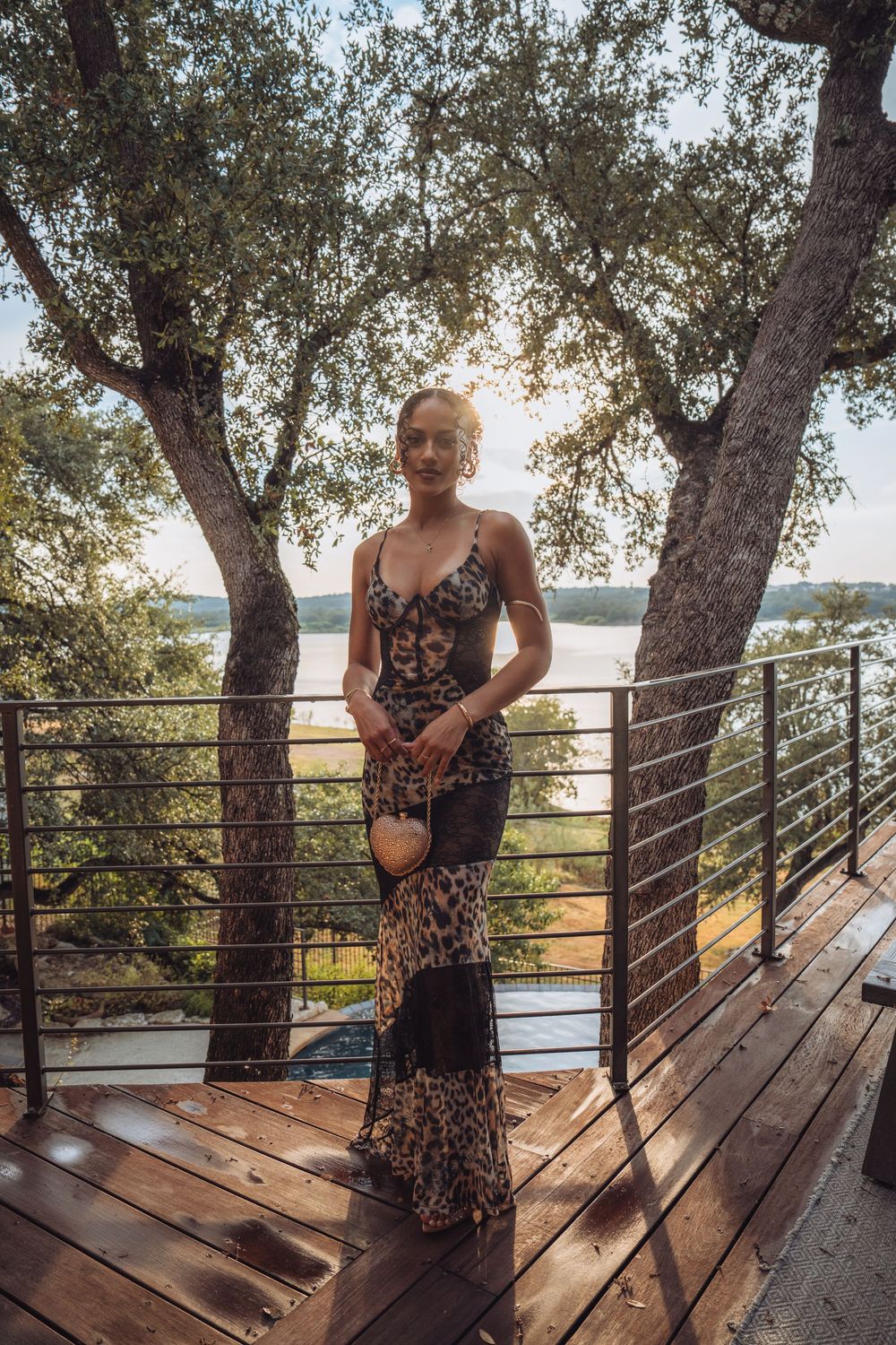 Woman in leopard print maxi dress standing on wooden deck surrounded by trees with lake view at sunset.