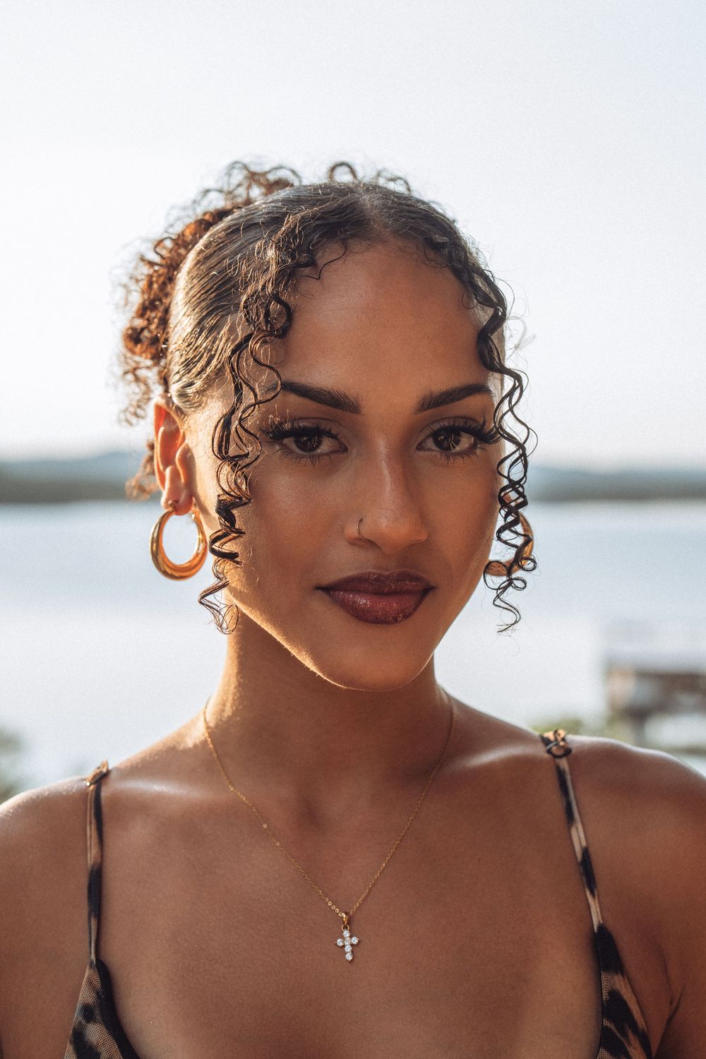 Young woman with curly hair wearing hoop earrings and a cross necklace poses outdoors near water.