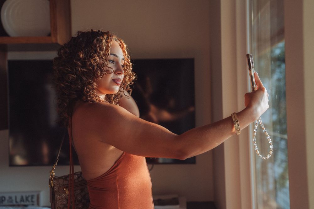 A curly-haired woman in an orange dress takes a selfie by a sunlit window wearing pearl bracelet and carrying a brown bag.