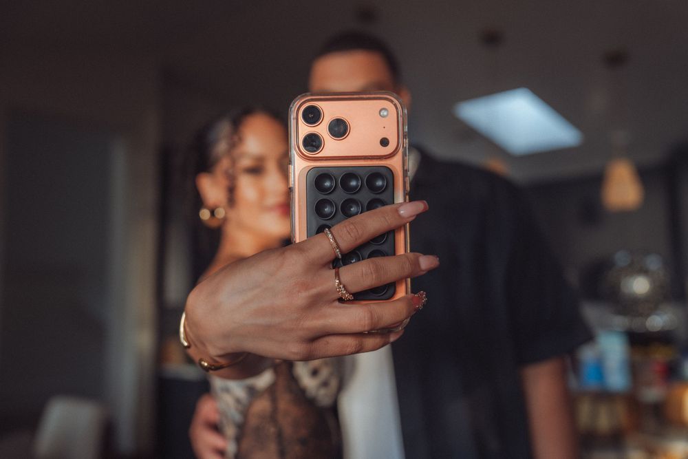 Couple taking a mirror selfie with a rose gold iPhone case featuring multiple camera lenses in a modern home.