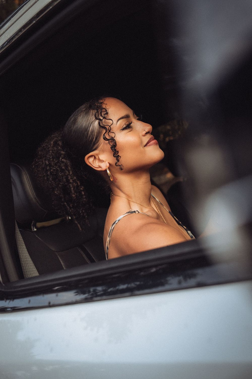 A young woman with curly hair sits in a car, eyes closed, wearing silver jewelry, bathed in warm sunlight.