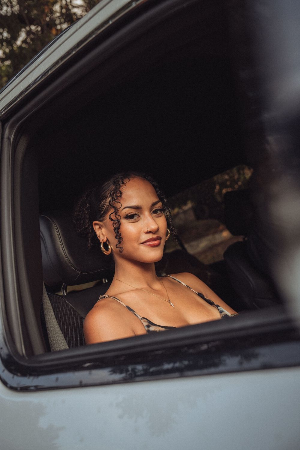 Young woman with curly hair and hoop earrings smiling inside a vehicle, viewed through an open car window.