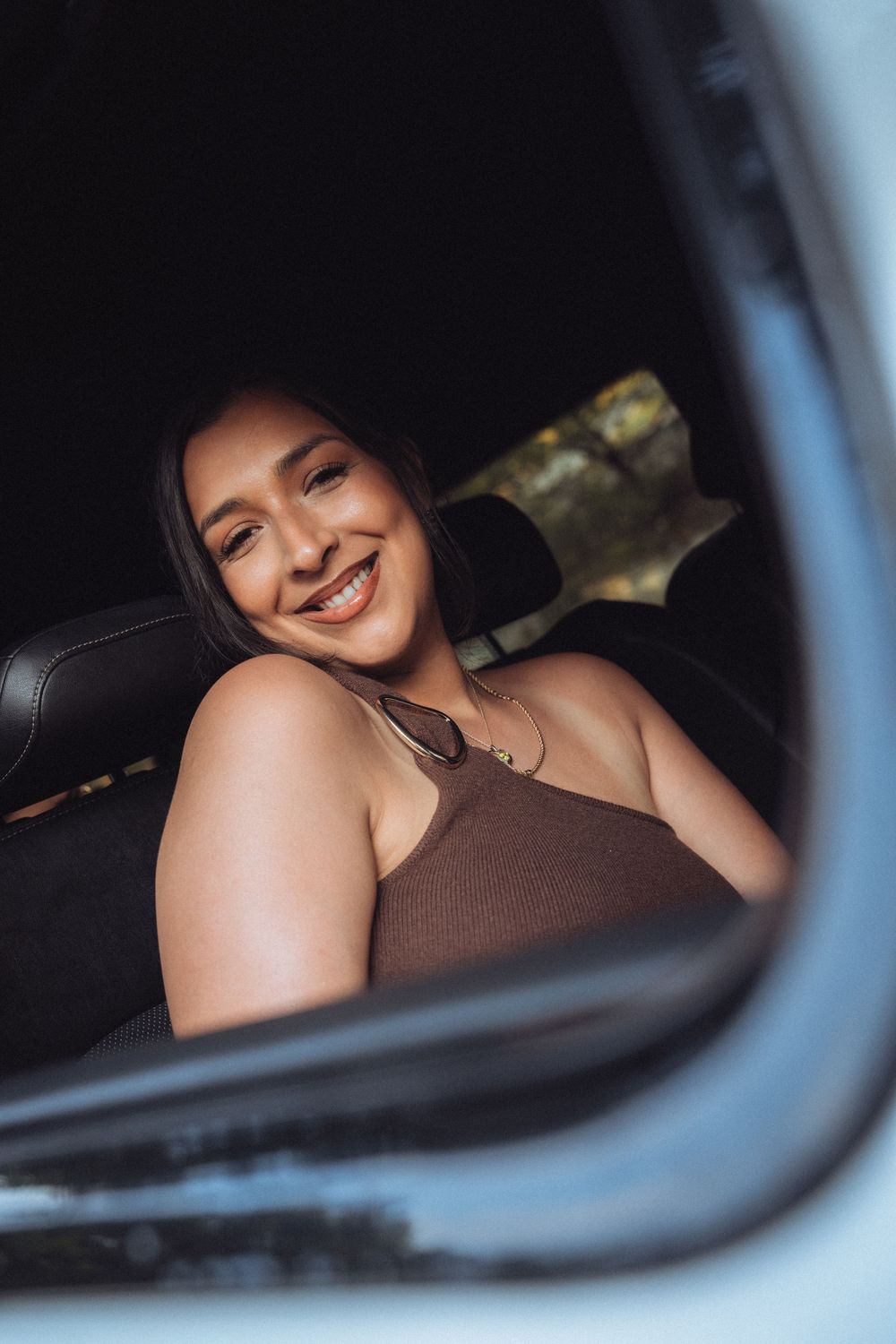 Smiling young woman in brown tank top sitting in car backseat, viewed through window reflection.