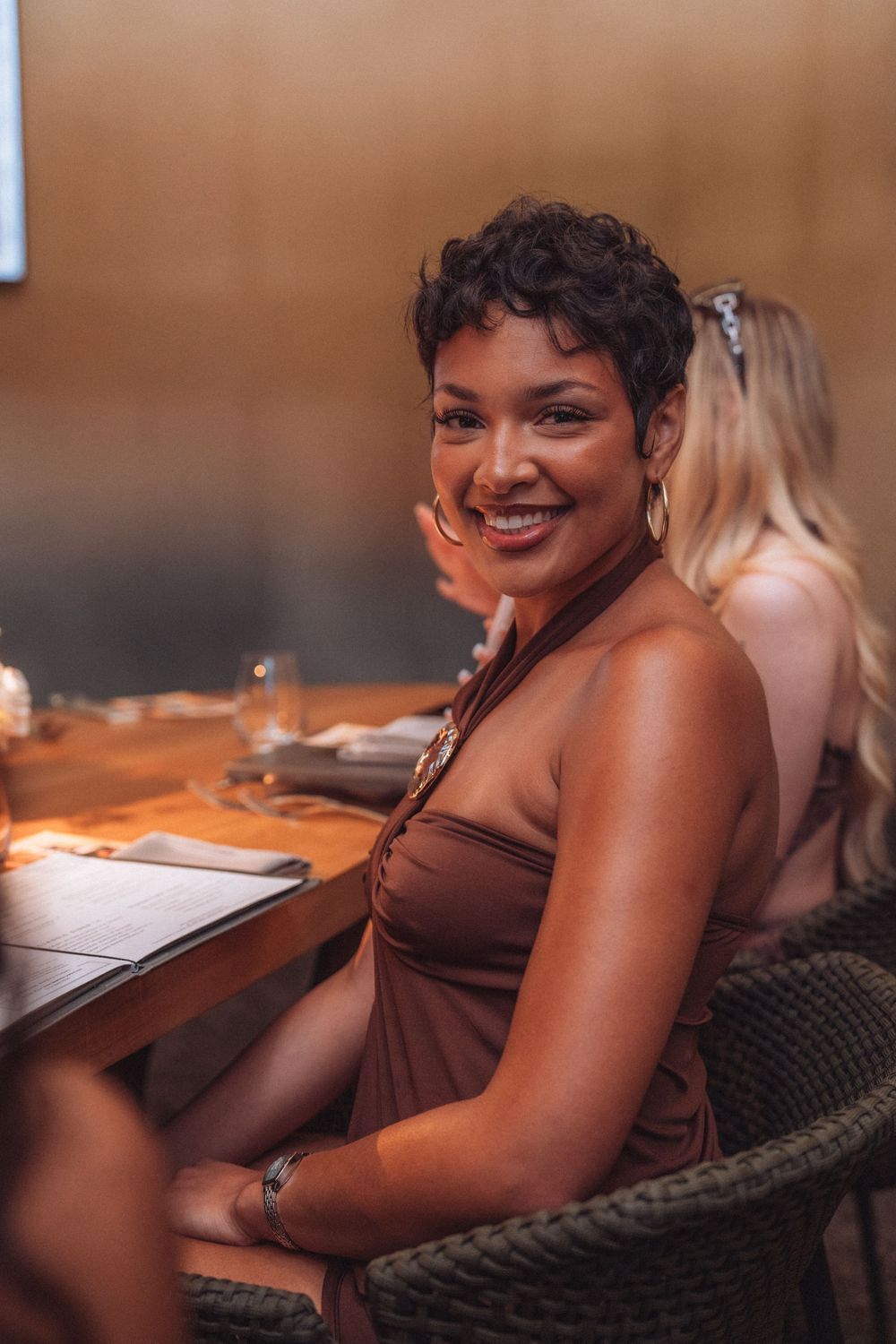 Smiling woman in brown halter dress seated at restaurant table with wine glasses, enjoying a night out with friends.