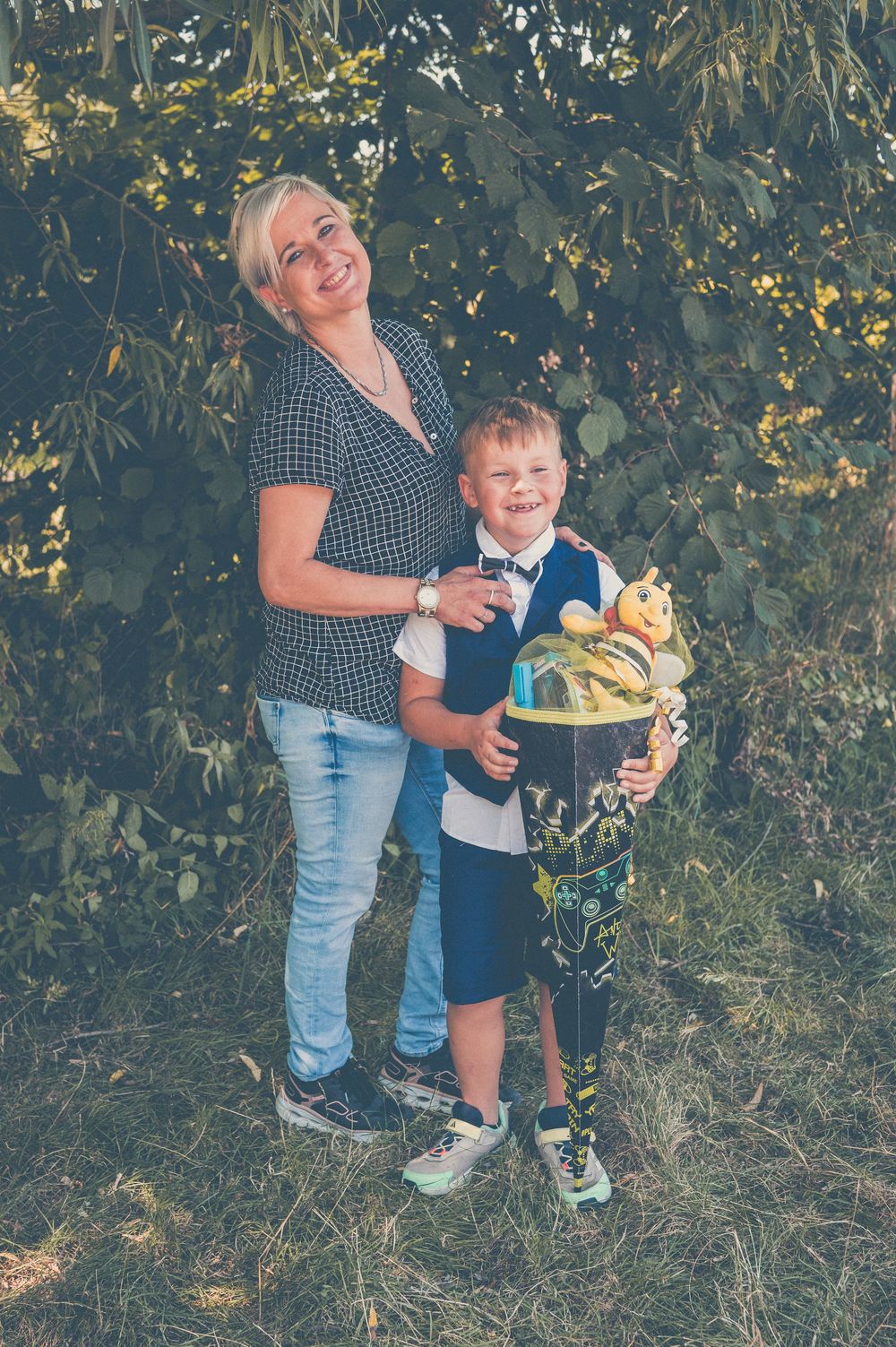 A smiling blonde woman and young boy in formal attire hold a decorative gift basket outdoors near green trees.