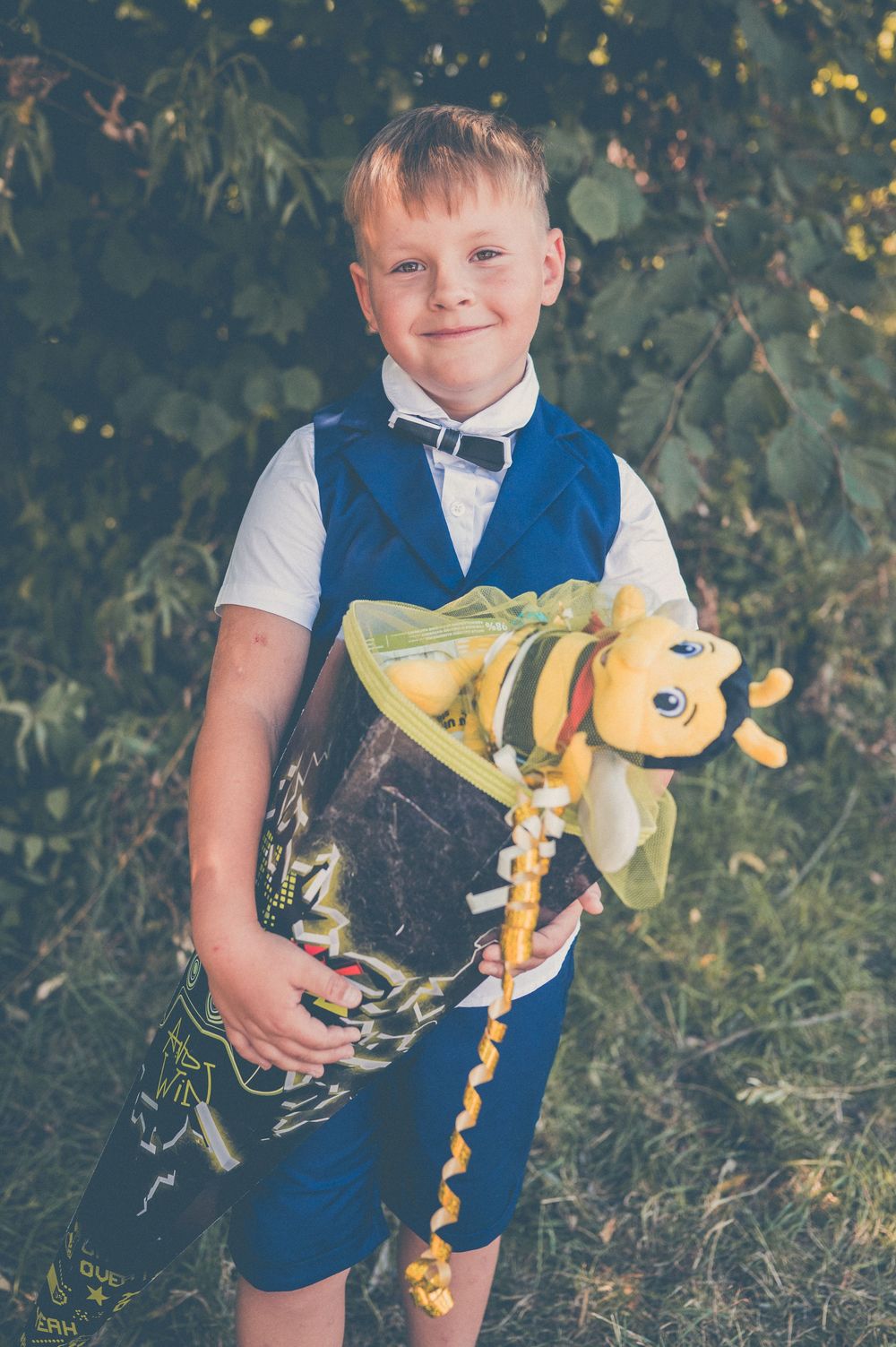 A smiling young boy in a blue vest and bow tie holds a yellow bee stuffed animal outdoors near green foliage.