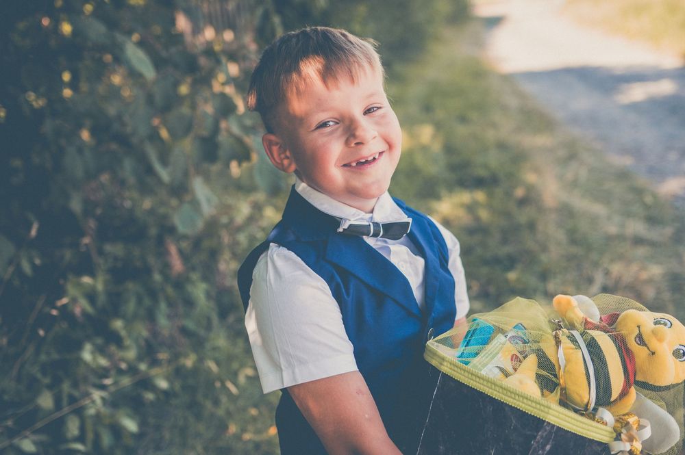 Smiling boy in blue vest and bow tie holding a yellow tray with toys outdoors.