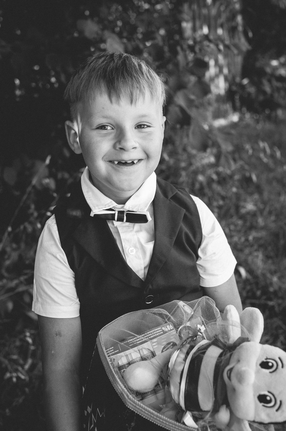 Smiling young boy in vest and bow tie holds a lunch bag with a stuffed bee toy, black and white portrait photo.