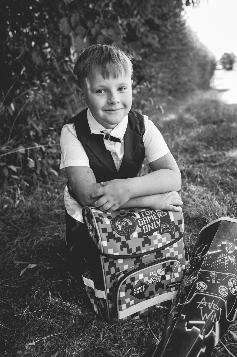 Black and white photo of a smiling boy in school uniform sitting outdoors with a gamers backpack.