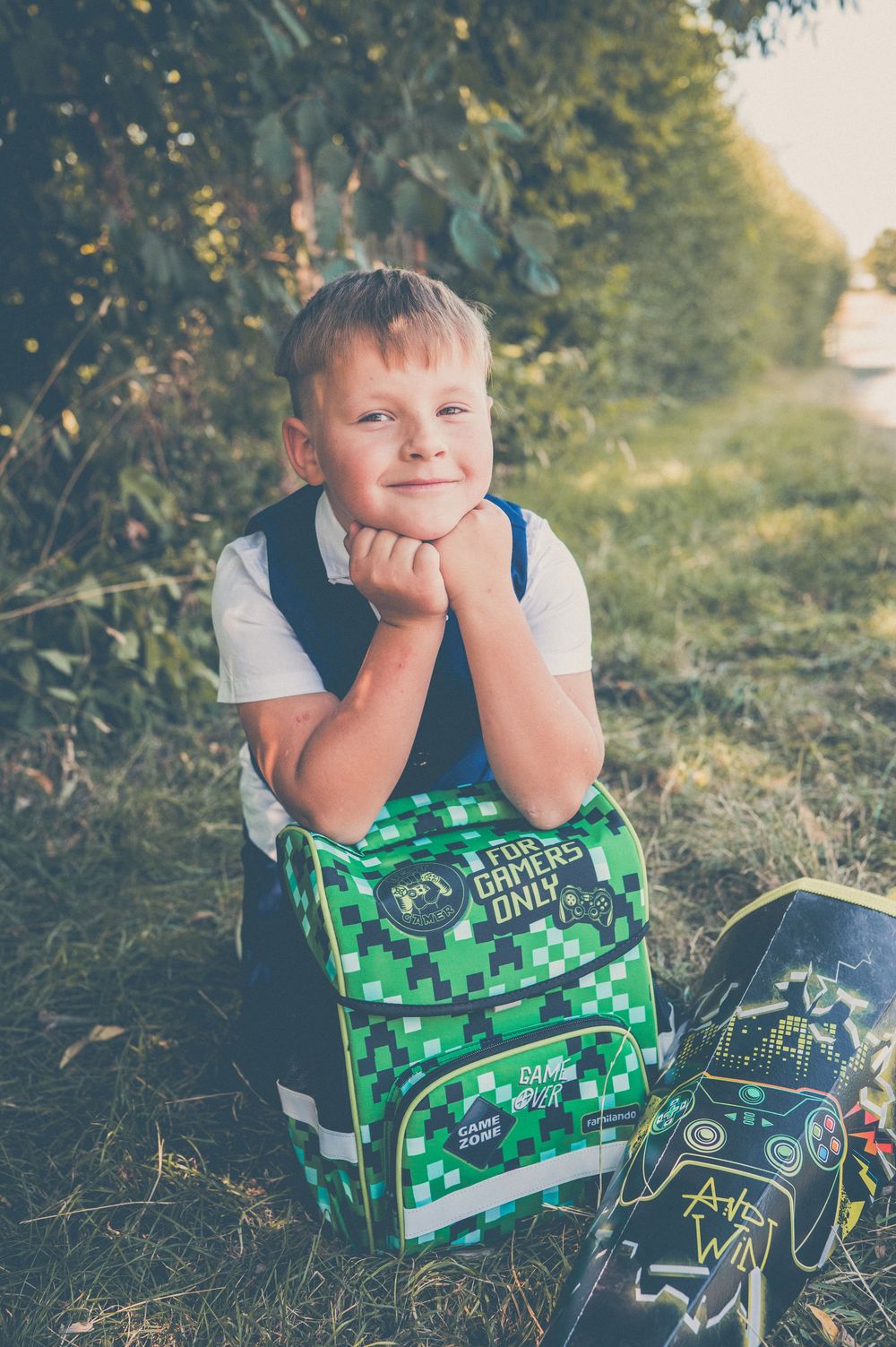 A smiling boy rests his chin on a green Minecraft-themed school backpack while sitting outdoors on grass.