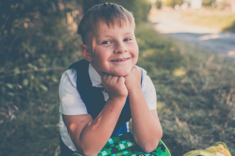 Smiling young boy with blue eyes resting chin on hands, sitting outdoors in natural light.
